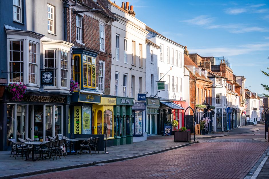 A wide street view in Sutton with a mixture of small retail shops and service outlets, including a pharmacy with a red sign, situated on either side of the pedestrianised high street. Several parked v
